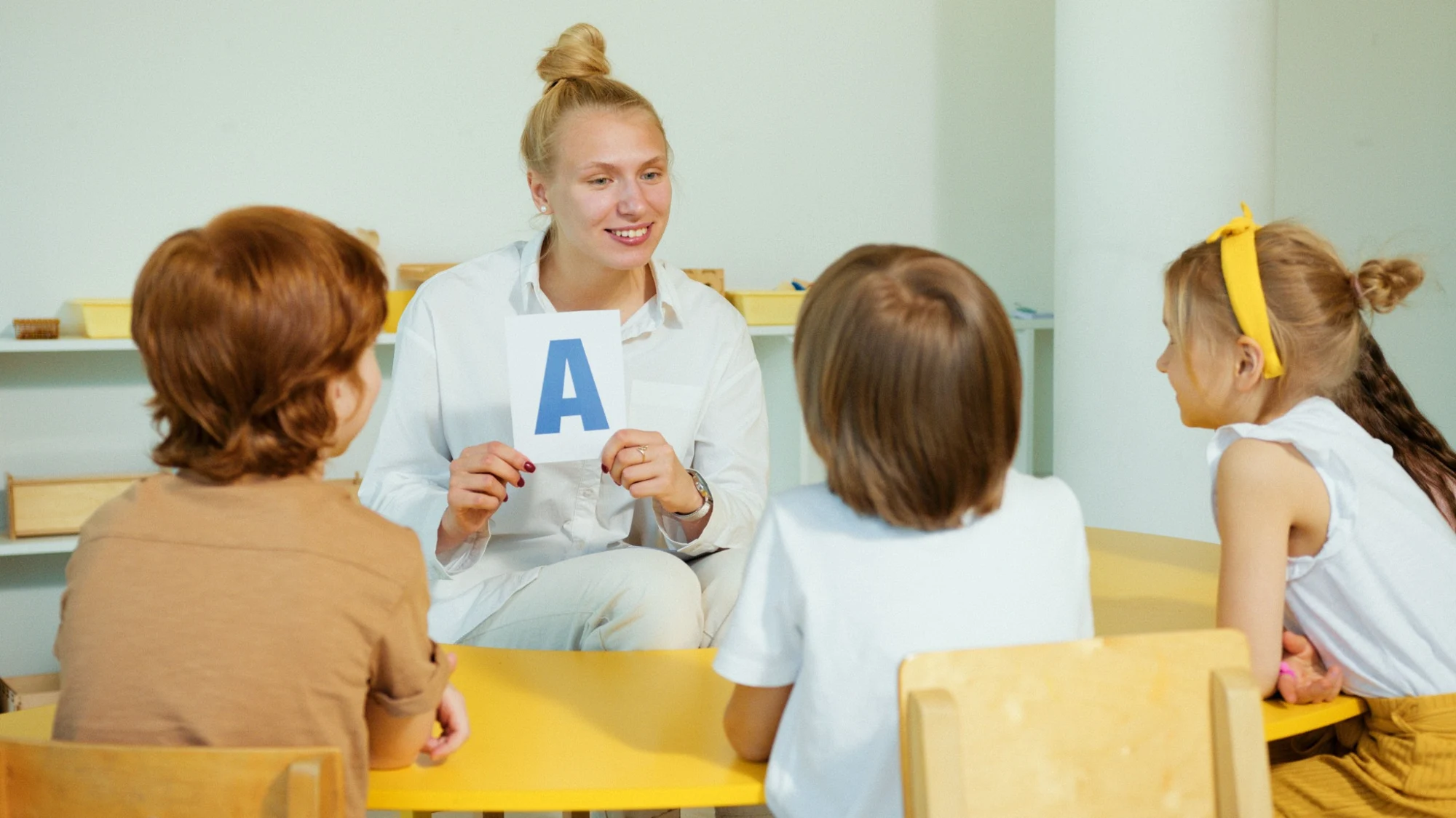 Lektorka pokazuje kartę z literą dzieciom przy żółtym stoliku